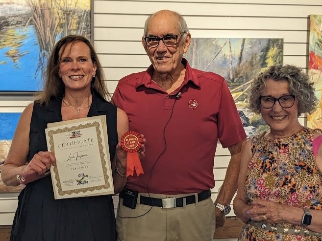 Three people smiling, holding a certificate and ribbon at an art award ceremony.
