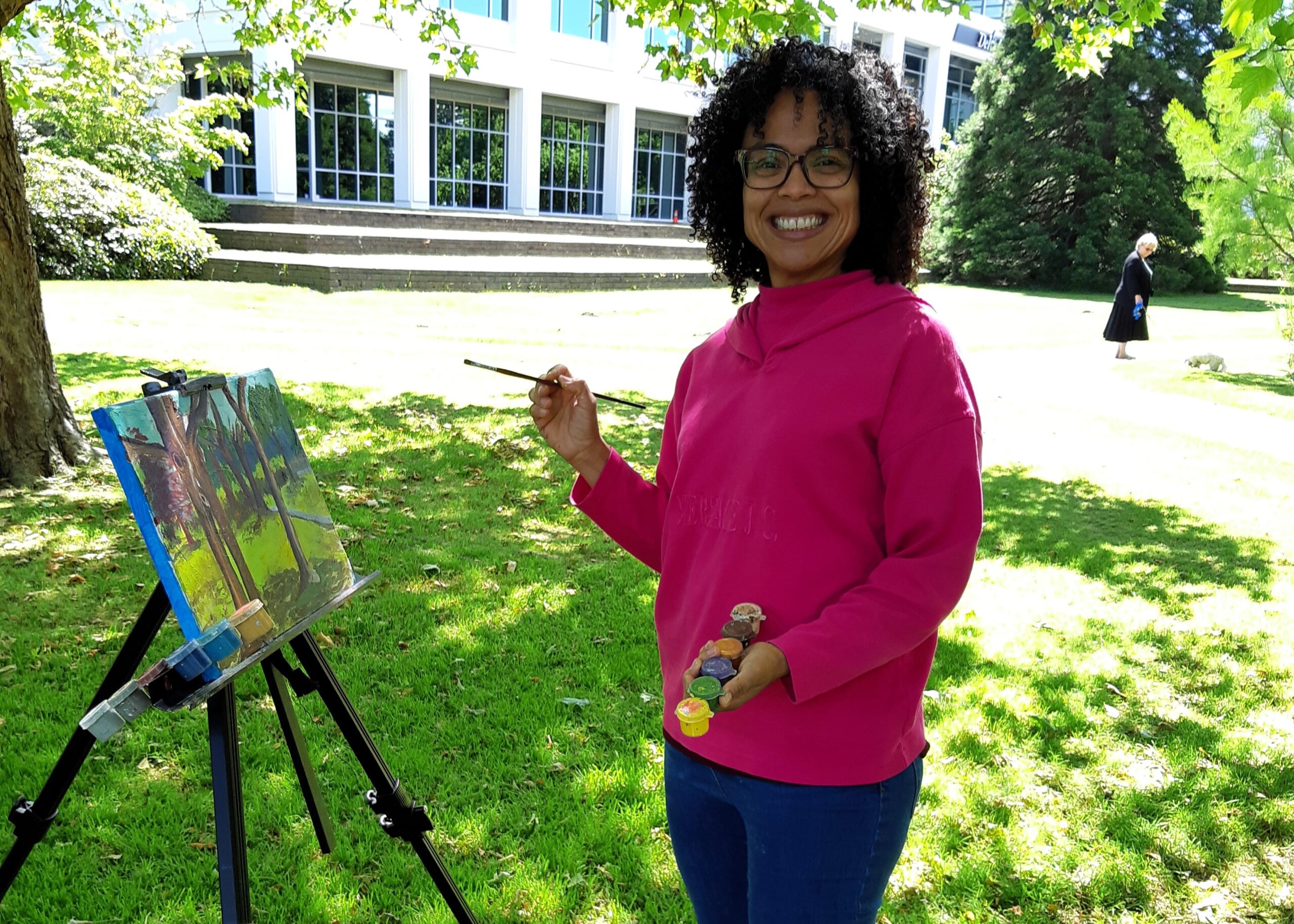 Artist painting outdoors on an easel while smiling at the camera.