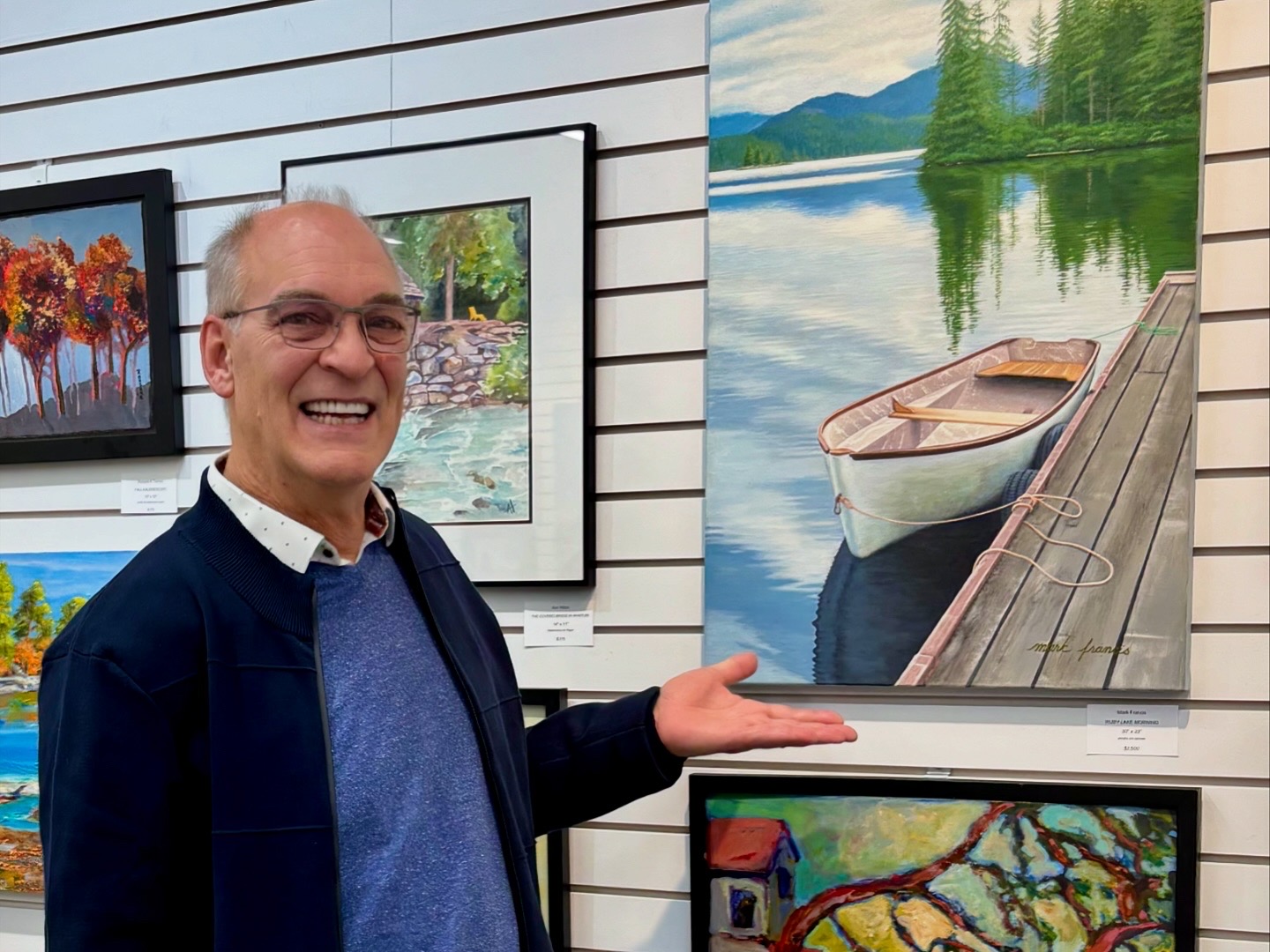 Man standing in front of a boat painting, smiling and gesturing proudly.